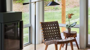 Ein modernes Zimmer mit großen Fenstern, einer Stehlampe, einem Fernseher, einem gewebten Sessel und einem kleinen Tisch mit einer Pflanze.
