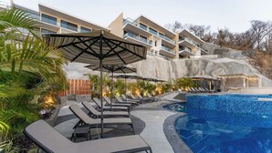 Een zwembad met ligbedden en parasols, een modern gebouw met balkons, en een heuvellandschap op de achtergrond.