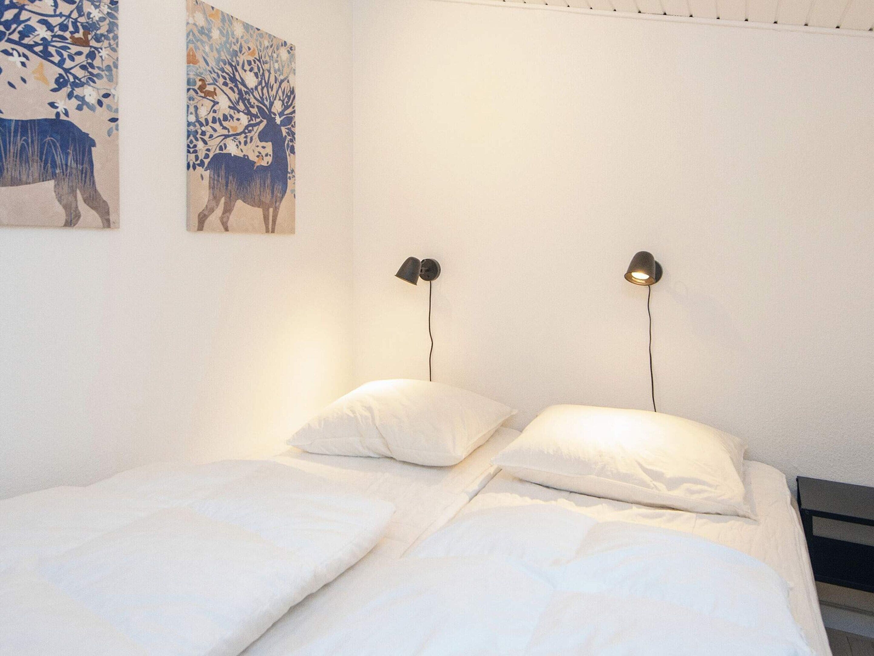 Una cama con ropa blanca, dos lámparas negras fijadas a la pared y dos cuadros colgados.