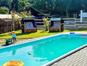 Ein Schwimmbecken mit Liegestühlen, eine Person mit einem Schubkarren und ein eingezäunter Bereich mit Bäumen.
