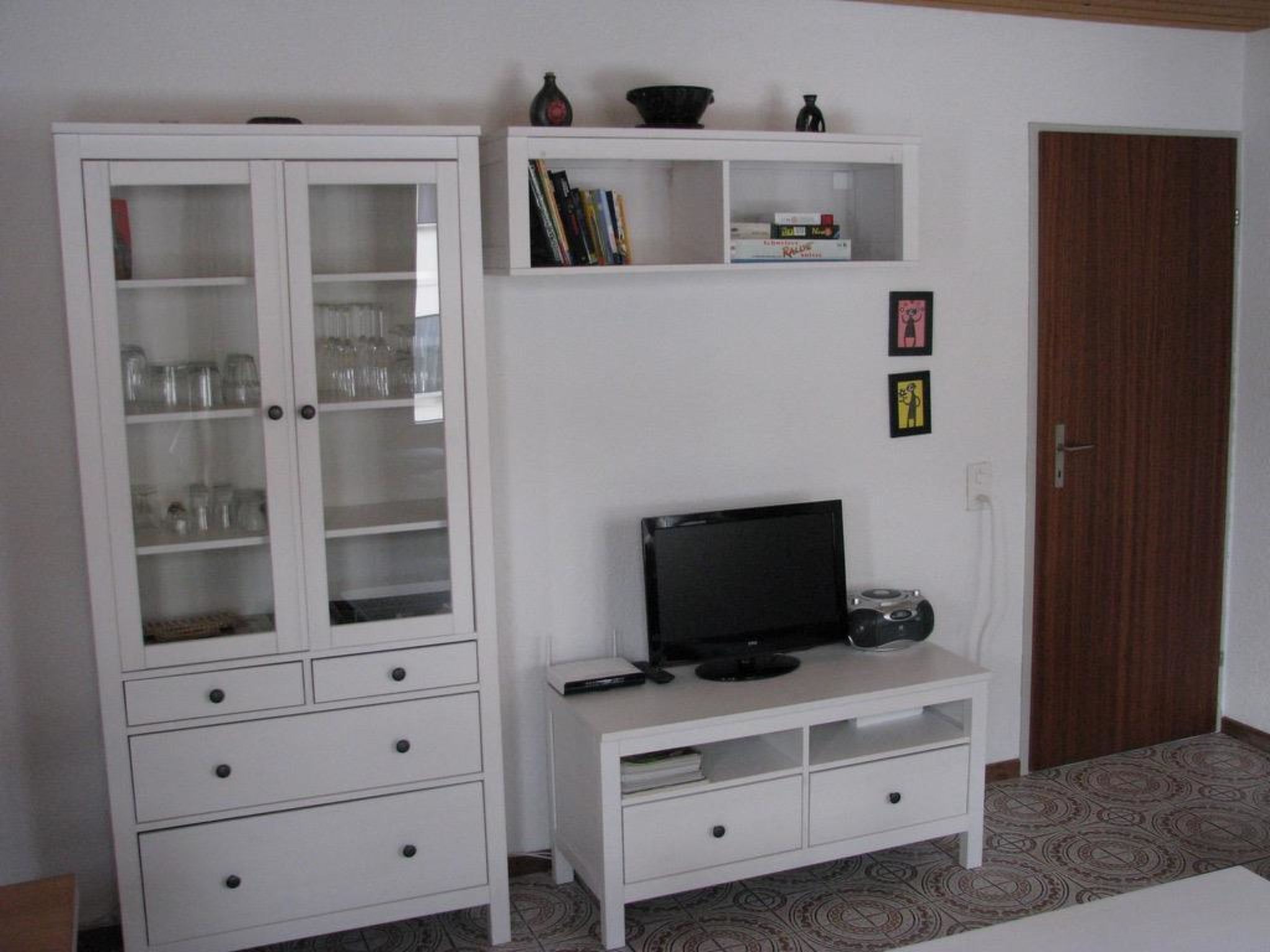 Une pièce avec un meuble blanc doté de portes en verre, un meuble de rangement blanc contenant une télévision et une radio, et un tapis à motifs sur le sol.