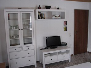 Ein Zimmer mit einem weißen Schrank mit Glastüren, einer weißen Unterhaltungsanlage inklusive Fernseher und Radio sowie einem gemusterten Teppichboden.