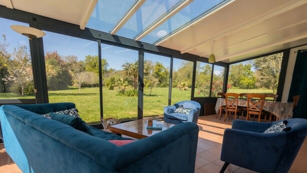 Een moderne woonkamer met een glazen dak, een blauwe bank, een kleine tafel en een eethoek met stoelen.