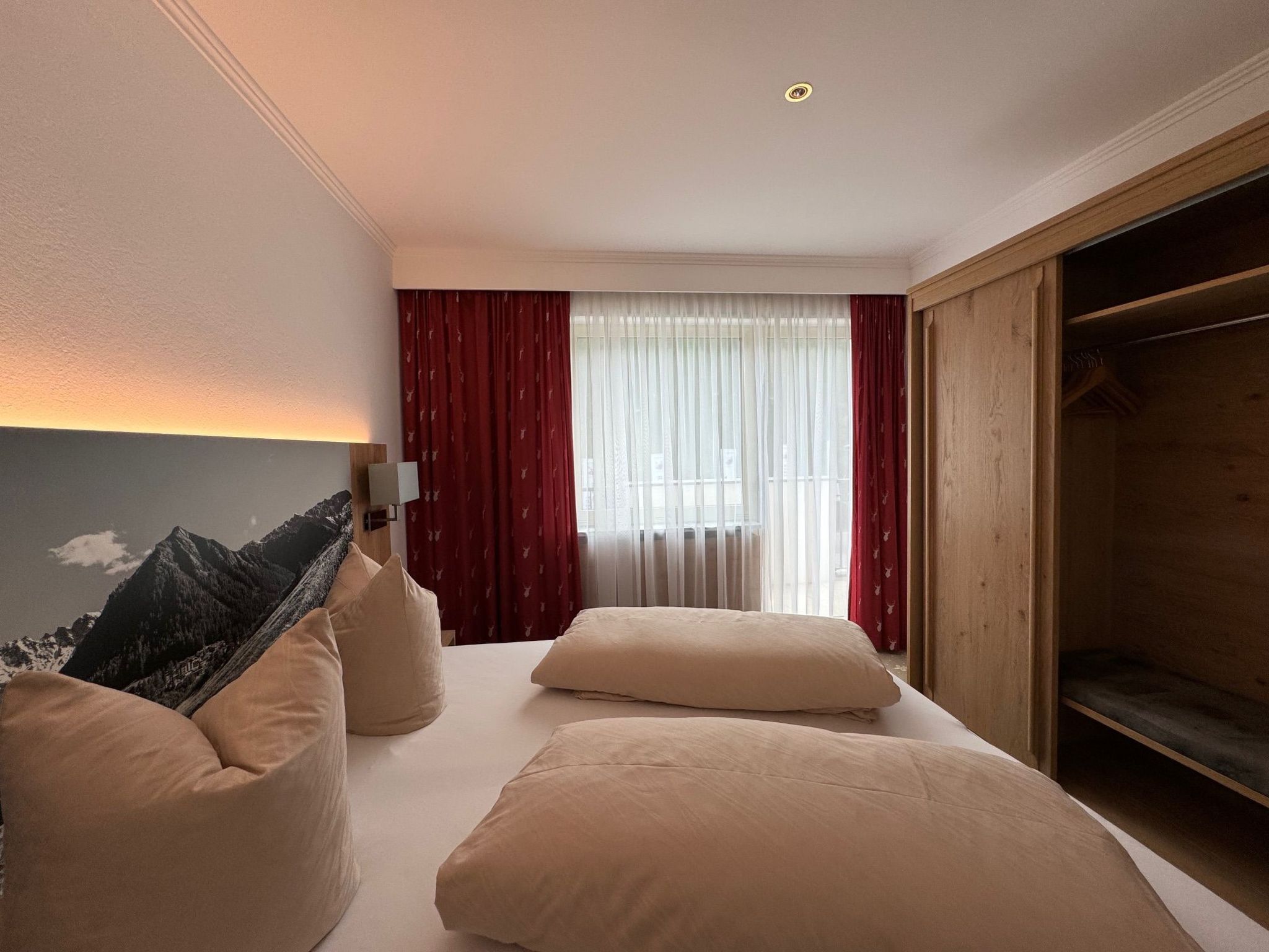 Una camera d'albergo con un letto, un comodino, una lampada, un armadio e vista sulle montagne dalla finestra.
