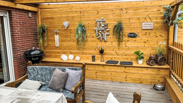Une terrasse en bois équipée d’un barbecue, d’un coin salon avec des coussins et de divers éléments décoratifs.