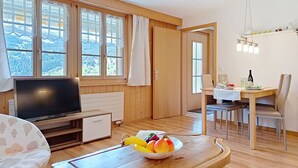 Een kamer met een televisie, een eettafel met stoelen, een schaal fruit en grote ramen met gordijnen.
