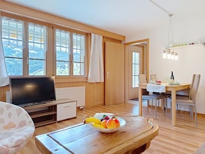 Ein Zimmer mit Fernseher, einem Esstisch mit Stühlen, einer Obstschale und großen Fenstern mit Vorhängen.