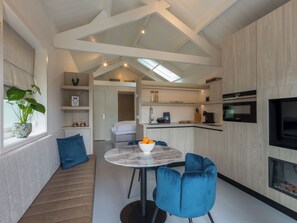 A modern kitchen with a built-in oven, a dining area with a round table and blue chairs, and a living area with a sofa and a plant. - Lepelaar by Interhome (Domburg)