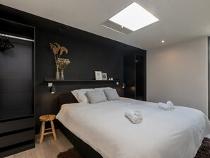 Ein modernes Schlafzimmer mit einem großen Bett, einem kleinen Hocker, einem Nachttisch mit Lampe und einem Wandregal mit Dekoration.