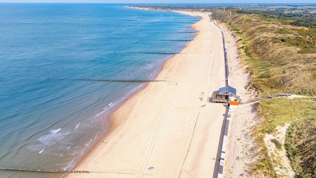 Une plage côtière avec une longue jetée qui s’avance dans l’eau, un petit bâtiment doté de panneaux solaires et un paysage vallonné en arrière-plan.