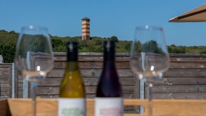 Eine Holzterrasse im Vordergrund mit zwei Weingläsern und zwei Weinflaschen, im Hintergrund ein Leuchtturm.