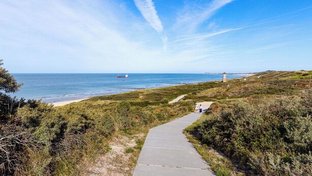 Ein Küstenpfad, der zu einem Strand führt, mit einem Leuchtturm in der Ferne.