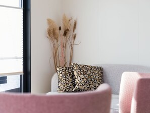 Een kamer met een grijze bank, een roze stoel en een decoratieve pampasgrasopstelling tegen een witte muur.