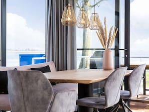 Een moderne eetruimte met een houten tafel, grijs gestoffeerde stoelen en uitzicht op de zee door grote ramen.