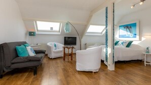 Ein geräumiger Raum mit Holzboden, einem grauen Sofa, einem weißen Sessel, einem kleinen Holztisch, einem Fernseher und einem Bett mit weißer Bettwäsche.