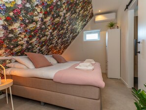 Ein Schlafzimmer mit schrägen Wänden, einem Bett mit Kissen und Decke, einem Nachttisch mit Lampe und einem Fenster.