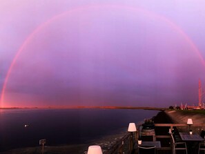 A sunset view with a rainbow over a calm body of water, wooden deck with tables and chairs, and a distant shoreline with structures. - Aurum Solis by Interhome (Sint-Annaland)