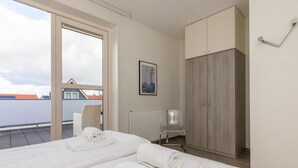 Ein Hotelzimmer mit zwei Betten, einem Heizkörper, einem Sessel, einem Kleiderschrank und einem Fenster mit Blick auf Gebäude.