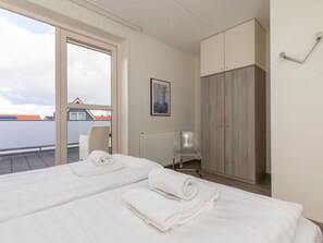 A hotel room with two beds, a radiator, a chair, a wardrobe, and a window with a view of buildings. - Zoete Licht by Interhome (Zoutelande)