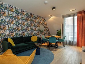 Ein modernes Wohnzimmer mit einem grünen Sofa, einem gelben Sessel und türkisfarbenen Stühlen. Ein runder Tisch mit Schale und Pflanze steht in der Mitte.