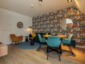Ein modernes Wohnzimmer mit einem runden Holztisch, türkisfarbenen Stühlen, einem grünen Sofa und floraler Tapete.
