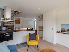 Een moderne keuken met witte kasten, een inbouwoven en een koffieapparaat. In de woonruimte staan een gele stoel en een blauwe bank.