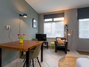 A modern living room with a wooden dining table, a TV, a sofa, and a chair. There are windows with curtains, a wall-mounted lamp, and a small rug on the floor. - Studio Rustenpolder by Interhome (Vrouwenpolder)