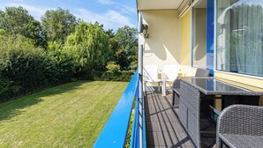 Un balcón con vistas a un área verde y árboles, una mesa con sillas y una ventana con cortinas.