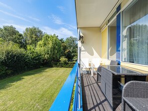 Een balkon met uitzicht op een gazon en bomen, een tafel met stoelen, en een raam met gordijnen.
