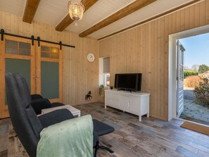 Ein Zimmer mit Holzwänden, einem Deckenventilator, einem Fernseher auf einem weißen Schrank, einer Uhr und einer Schiebetür, die nach draußen führt.