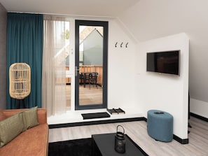 A modern living room with a sofa, a TV, a wicker floor lamp, a blue ottoman, and a sliding glass door leading to a balcony. - Bomont ZandStrand by Interhome (Renesse)