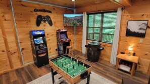 A room with wooden walls, a foosball table, two arcade machines, a window with a view, and a mounted game controller. - Bear Hug Hideaway: Secluded 2 King Cabin, Epic Arcade +Hot Tub, 4 min to Parkway (Sevierville)
