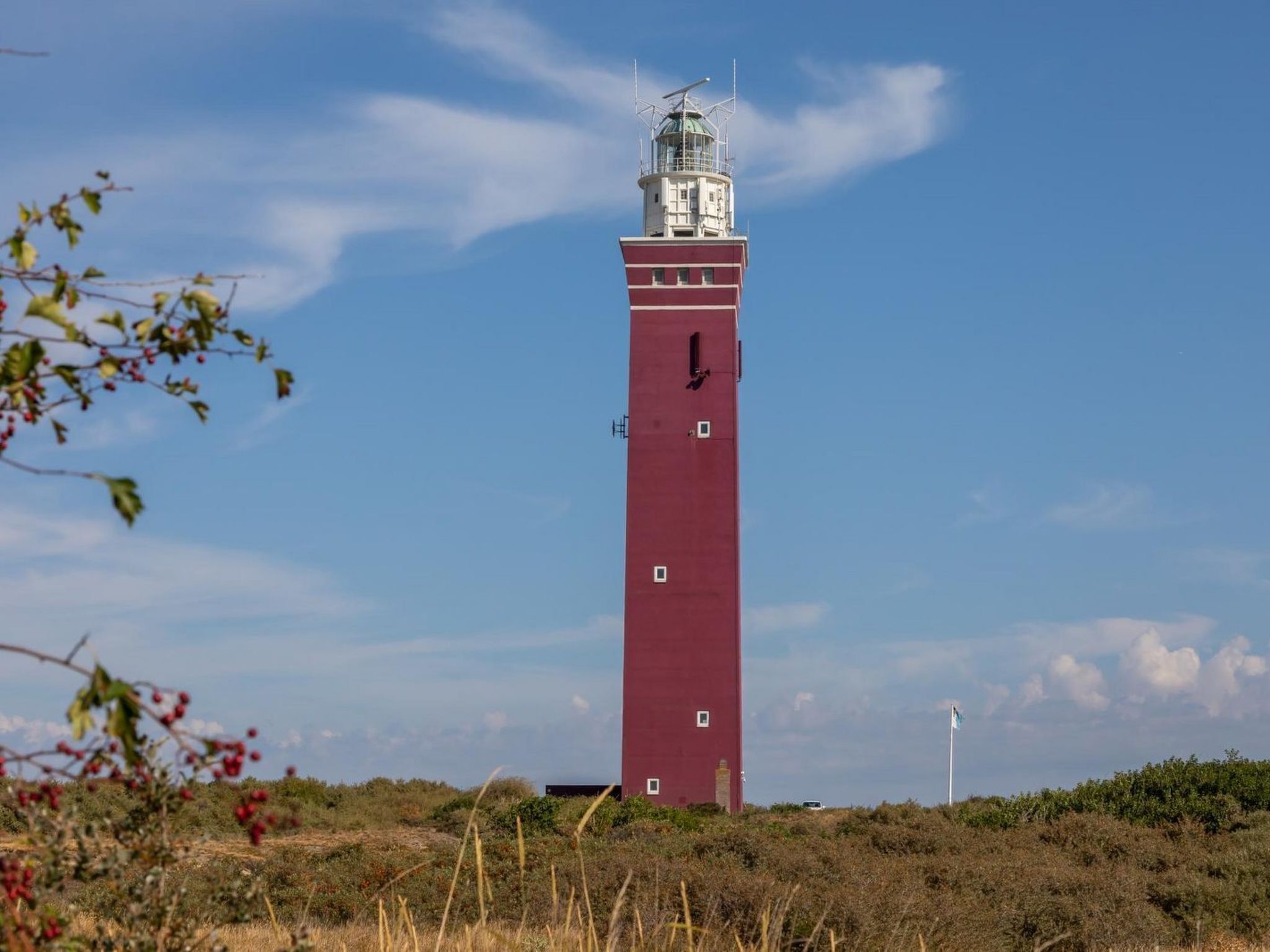 A tall, red lighthouse with a white top, surrounded by vegetation and a clear sky.