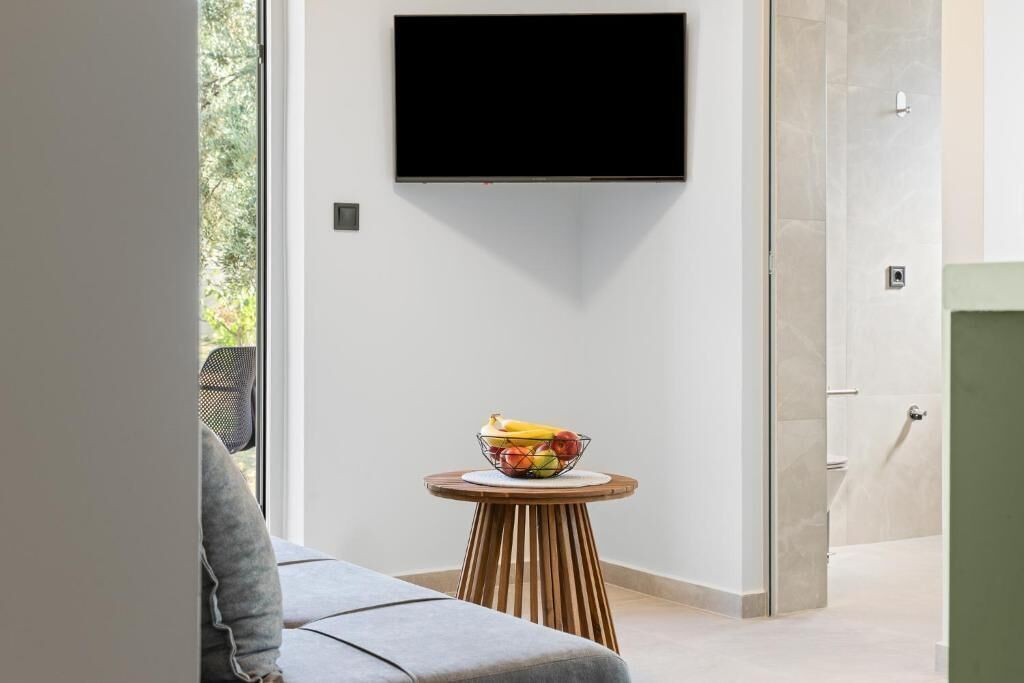 Ein modernes Zimmer mit einem an der Wand befestigten Flachbildfernseher, einem runden Holztisch mit Obstschale und einem grauen Sofa.