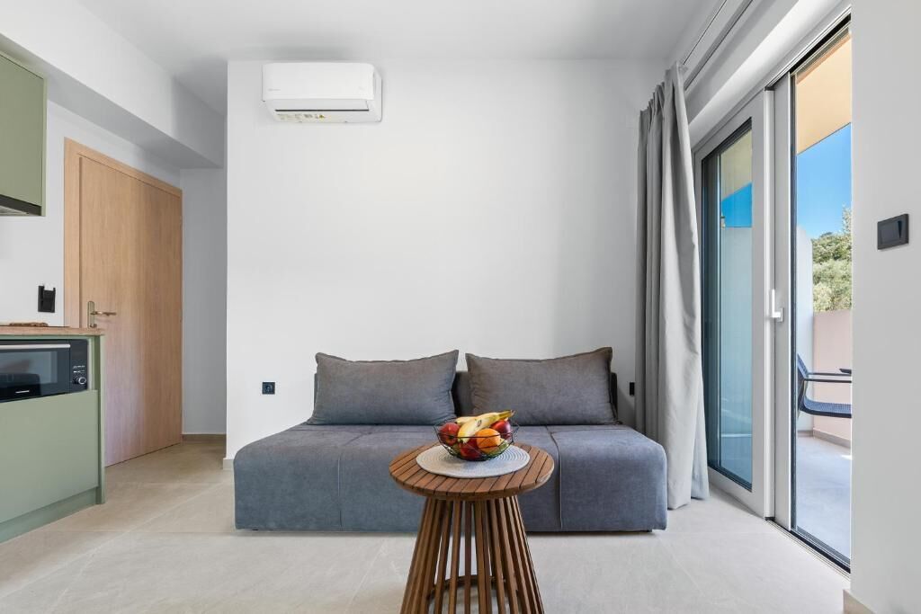 Ein modernes Zimmer mit einem grauen Sofa, einem runden Holztisch mit Obstschale, einer Küchenzeile und einer Schiebeglasdoor zum Außenbereich.