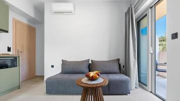 Ein modernes Zimmer mit einem grauen Sofa, einem runden Holztisch mit Obstschale, einer Küchenzeile und einer Schiebeglasdoor zum Außenbereich.
