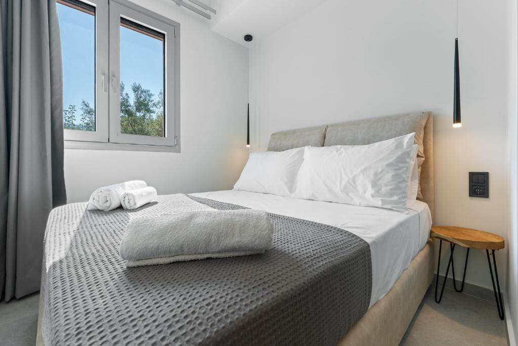 A modern hotel room with a large bed, a small bedside table, a window with curtains, and a wall-mounted light fixture.