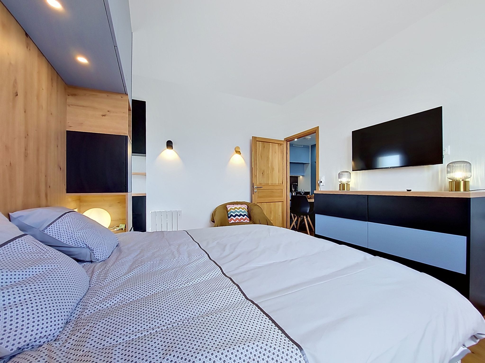 A modern bedroom with a large bed, a desk, a chair, and a TV mounted on the wall.