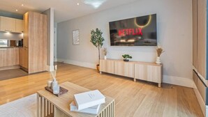 Ein modernes Wohnzimmer mit einem großen Flachbildfernseher, der 'NETFLIX' zeigt, einer Holzbank, einem Topf mit Pflanze und einem kleinen Tisch mit Büchern und einem Glas.