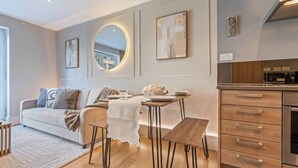 Ein modernes Wohnzimmer mit einer Couch, einem Esstisch mit Stühlen, einer Küchenzeile mit Schränken und einem großen Spiegel an der Wand.