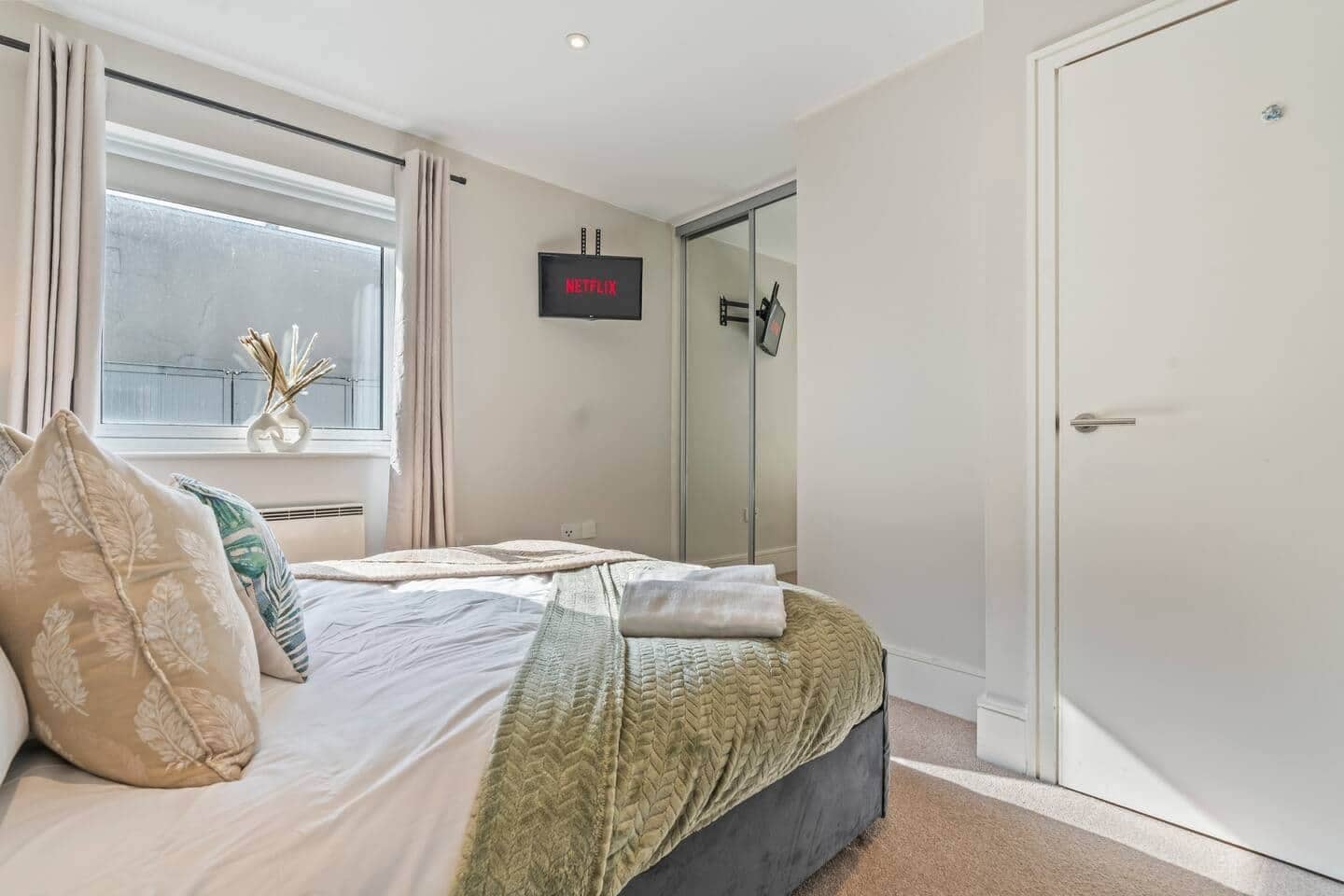 A bedroom with a bed, a window with curtains, a door, and a wall-mounted television displaying 'NETFLIX'.