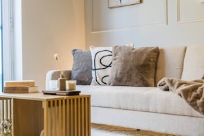 Una sala de estar moderna con un sofá, almohadones decorativos, una mesita con libros y una vela, y una ventana con luz natural.