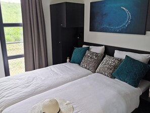 A double bed with white bedding, teal and gray patterned pillows, a black headboard, and a large window with curtains. - De Witte Parel by Interhome (Scherpenisse)