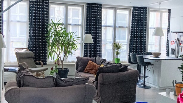 Een moderne woonkamer met een bank, fauteuils, een eethoek en grote ramen met gordijnen.