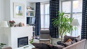 Een woonkamer met een bank, fauteuil en salontafel. Er is een boekenkast, een plant en een open haard.
