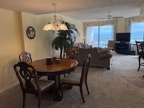 Ein Essbereich mit rundem Tisch und Stühlen, ein Wohnzimmer mit Sofa und Fernseher, eine große Pflanze und ein Balkon mit Aussicht.