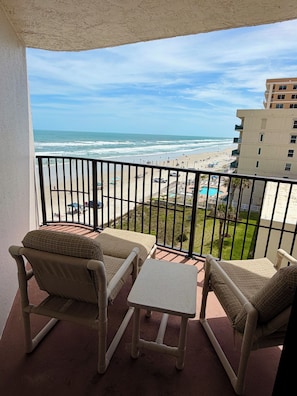Ein Balkon mit zwei Korbstühlen und einem kleinen Tisch, der auf einen Strand mit Pool und einem Gebäude im Hintergrund blickt.