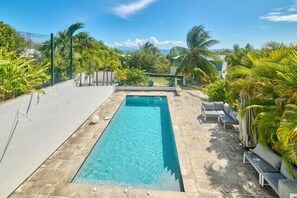 Ein rechteckiger Swimmingpool mit klarem Wasser, umgeben von Steinplatten, neben einer Liegefläche mit Bänken.