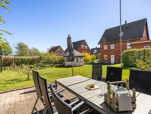 En udendørs terrasse med et bord og stole, en lille have og et rødstenshus i baggrunden.