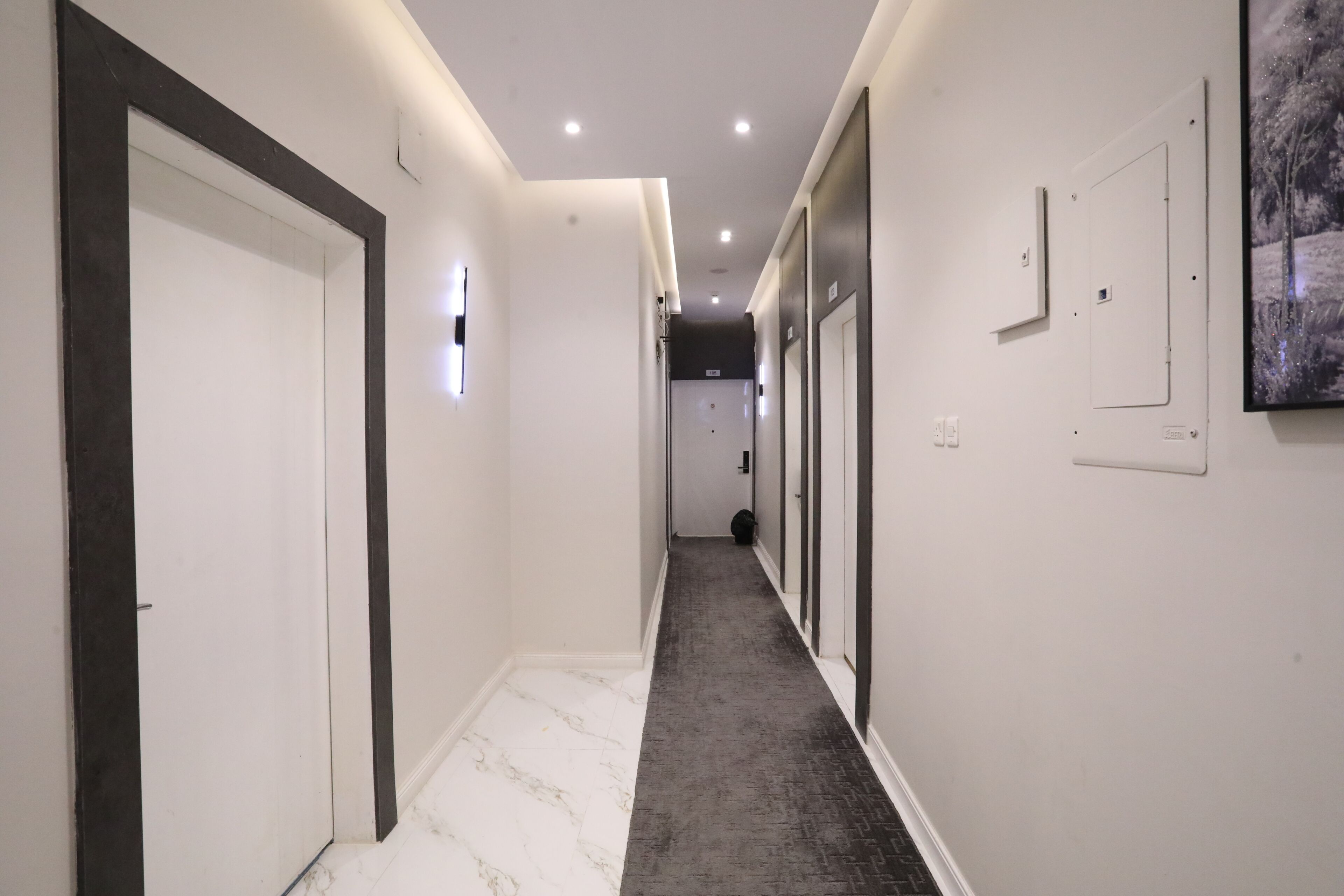 Un couloir aux murs blancs, aux moulures sombres et au sol en marbre, qui mène à une porte.